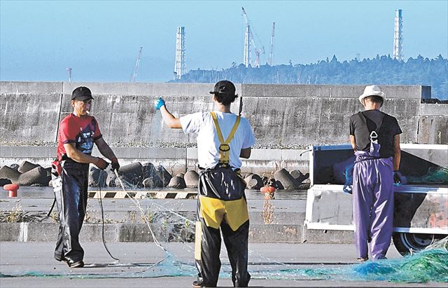 網の片付けをする漁師。奥は東京電力福島第1原発の排気筒=24日午前6時20分ごろ、浪江町・請戸漁港